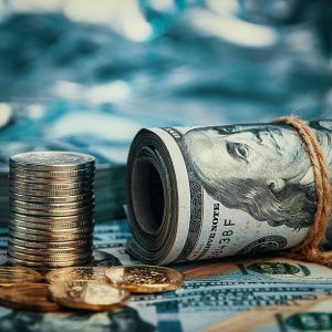 A roll of dollars with coins against a background of scattered hundred-dollar bills in a blue light. A wad of dollars, a roll of bills, and coins stacked and scattered.